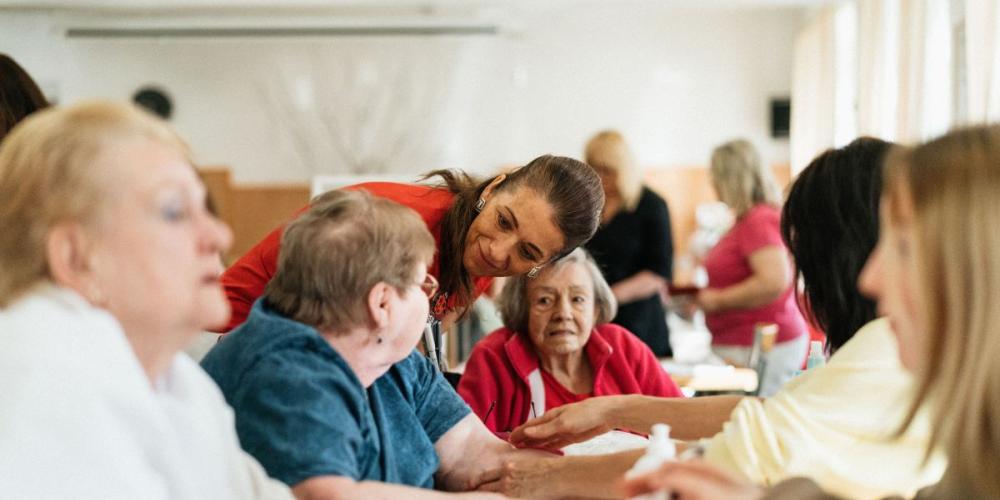 Zorgende dame spreekt iemand in groep oudere dames aan tafel aan
