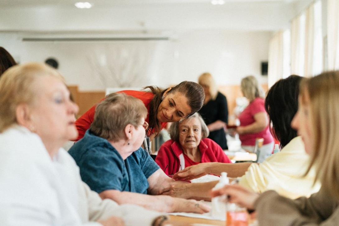 Zorgende dame spreekt iemand in groep oudere dames aan tafel aan